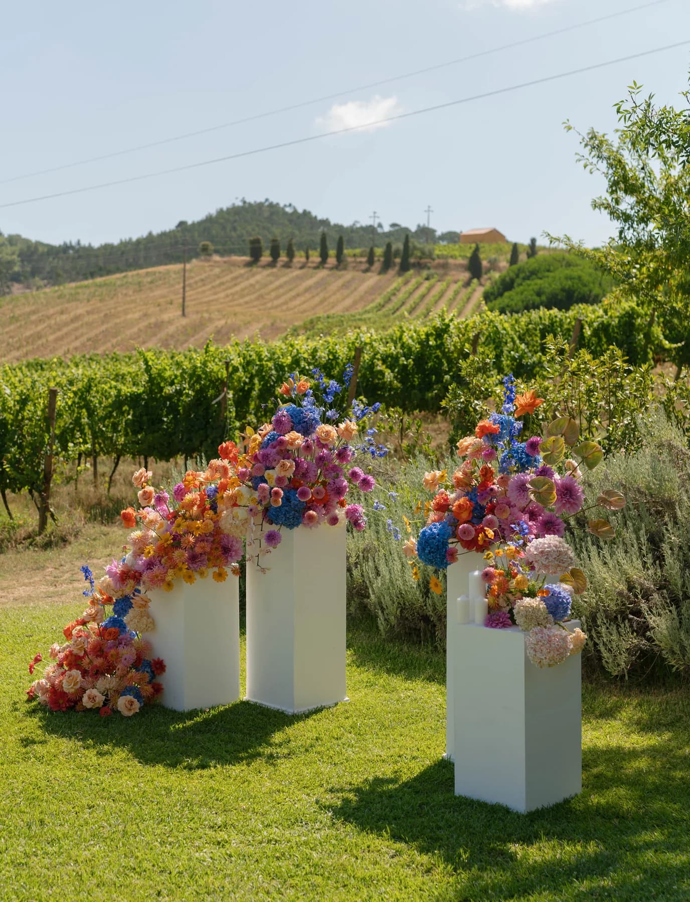 Bright ceremony flowers displayed on white plinths beside the vineyards