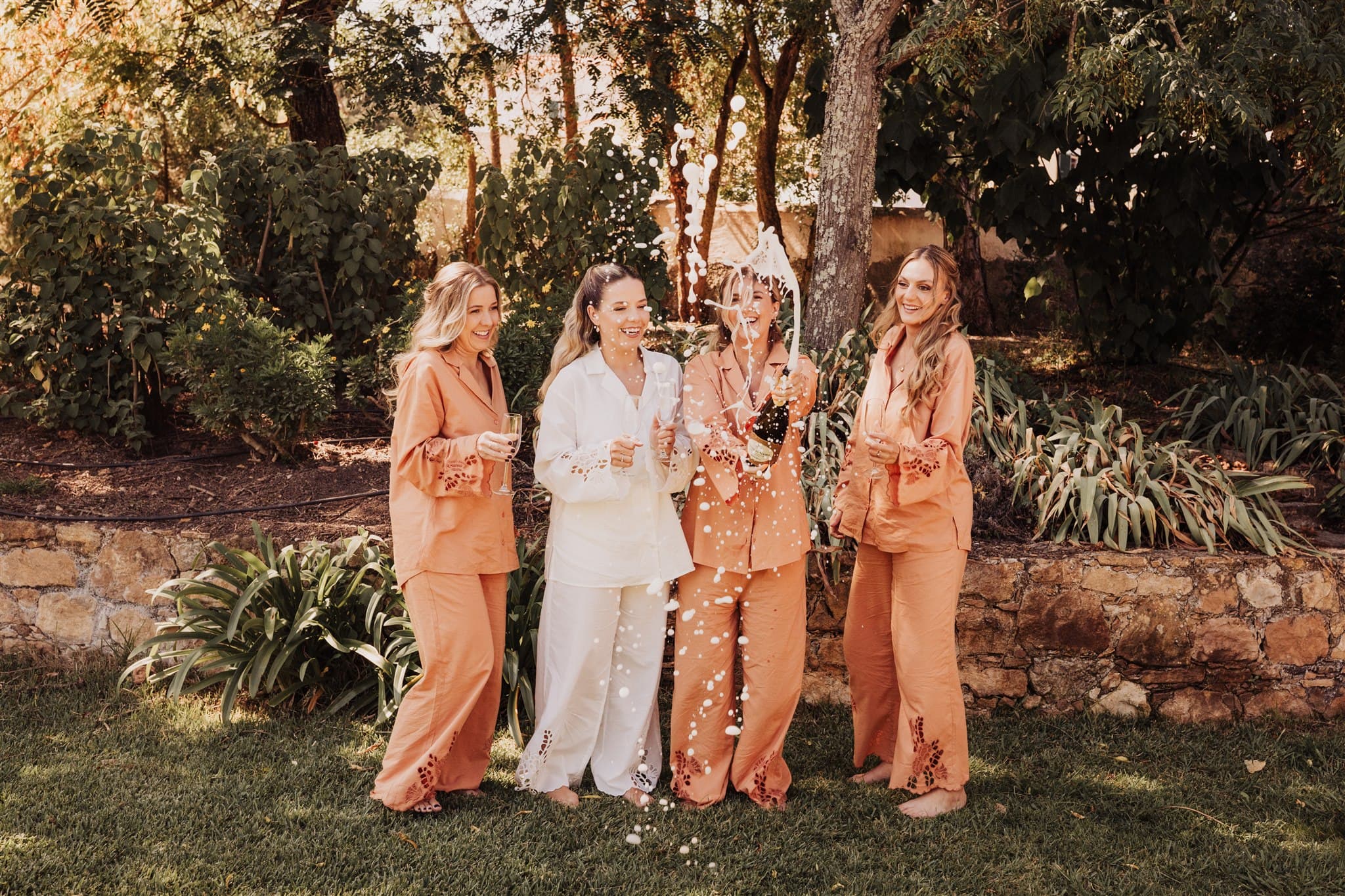 A bride and three friends in pyjamas laughing as they open sparkling wine in the Quinta's garden