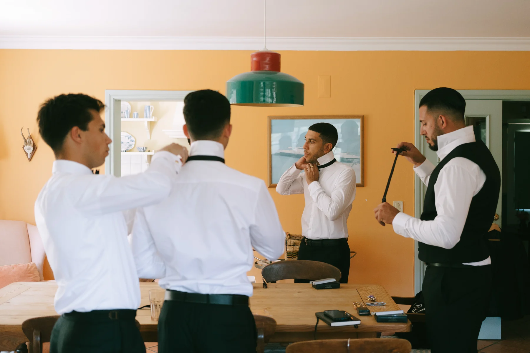Four men helping each other with their ties in a sunlit yellow room before the wedding